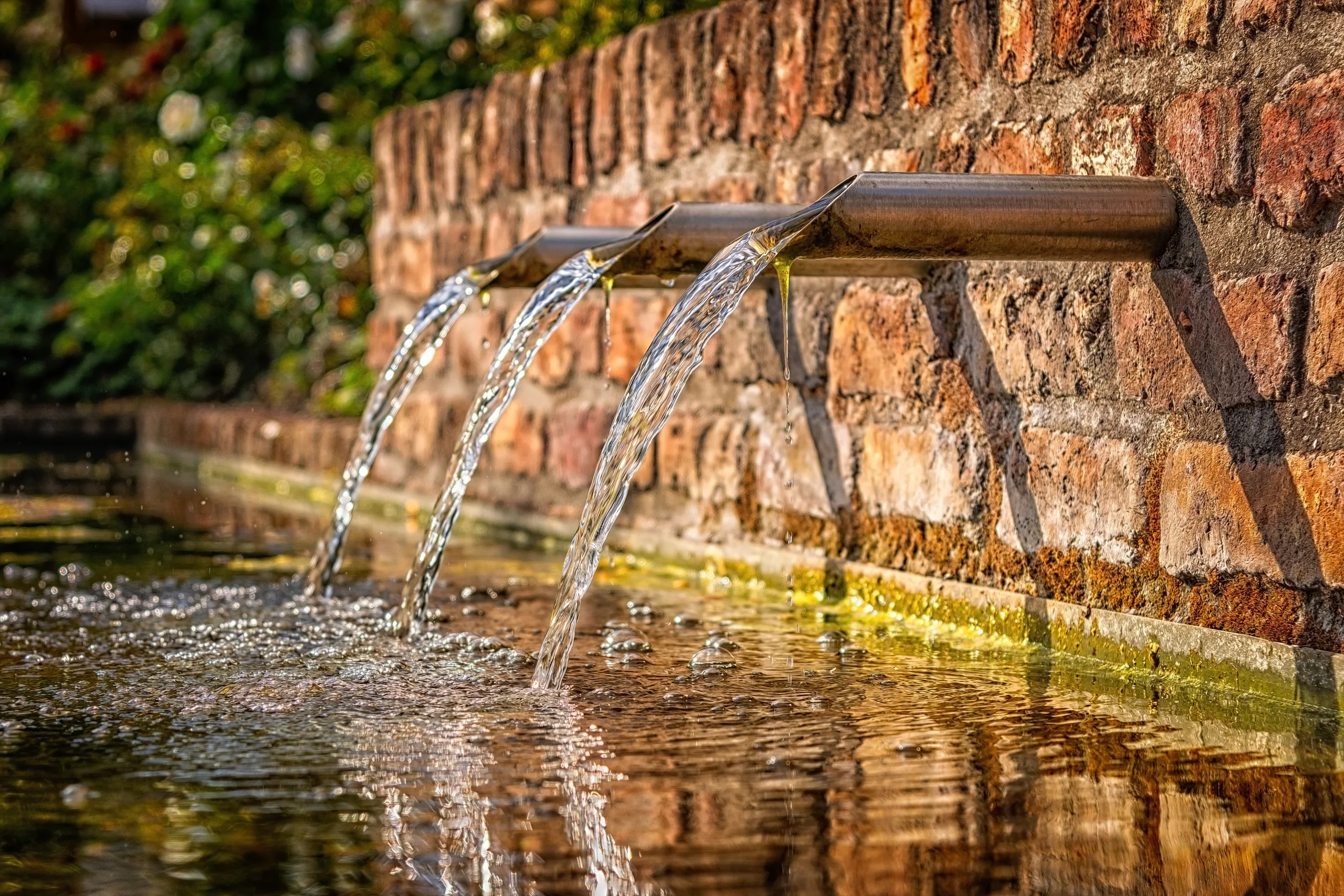 Three steel spouts pouring from a brick wall into a garden pond